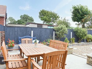 Une terrasse en bois avec une table et des chaises, entourée d’un petit muret en pierre et d’une haute clôture en bois.