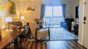 A modern living room with a wooden dining table, a sofa, and a chair. There is a balcony with a view of the outdoors. - Hillside Hideout - Great for groups! - Great Views (Branson)
