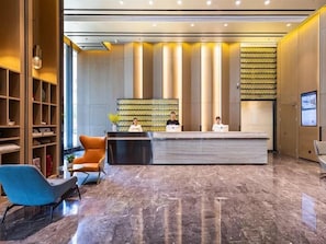 Ein Hotel-Rezeptionsbereich mit Empfangsschalter, zwei Mitarbeitern an der Rezeption, einem blauen Sessel und einem Bücherregal.