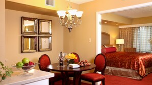 A hotel room with a dining area featuring a round table, chairs, and a fruit bowl. There is a chandelier above the table and a bed with a red bedspread in the background.