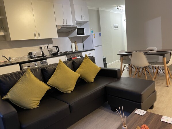 Una sala de estar moderna con un sofá seccional negro, una mesa de comedor con sillas y una cocina con armarios y electrodomésticos blancos.