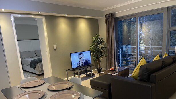 A modern living room with a dining area, a sofa, a TV, and a large window. - Tana’s Crib Modern Secure 1 bed apartment (Cape Town)
