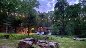 Una cabana rústica amb una fonda de pedra davant d'una zona boscosa, envoltada d'arbres i llums de garlandes.