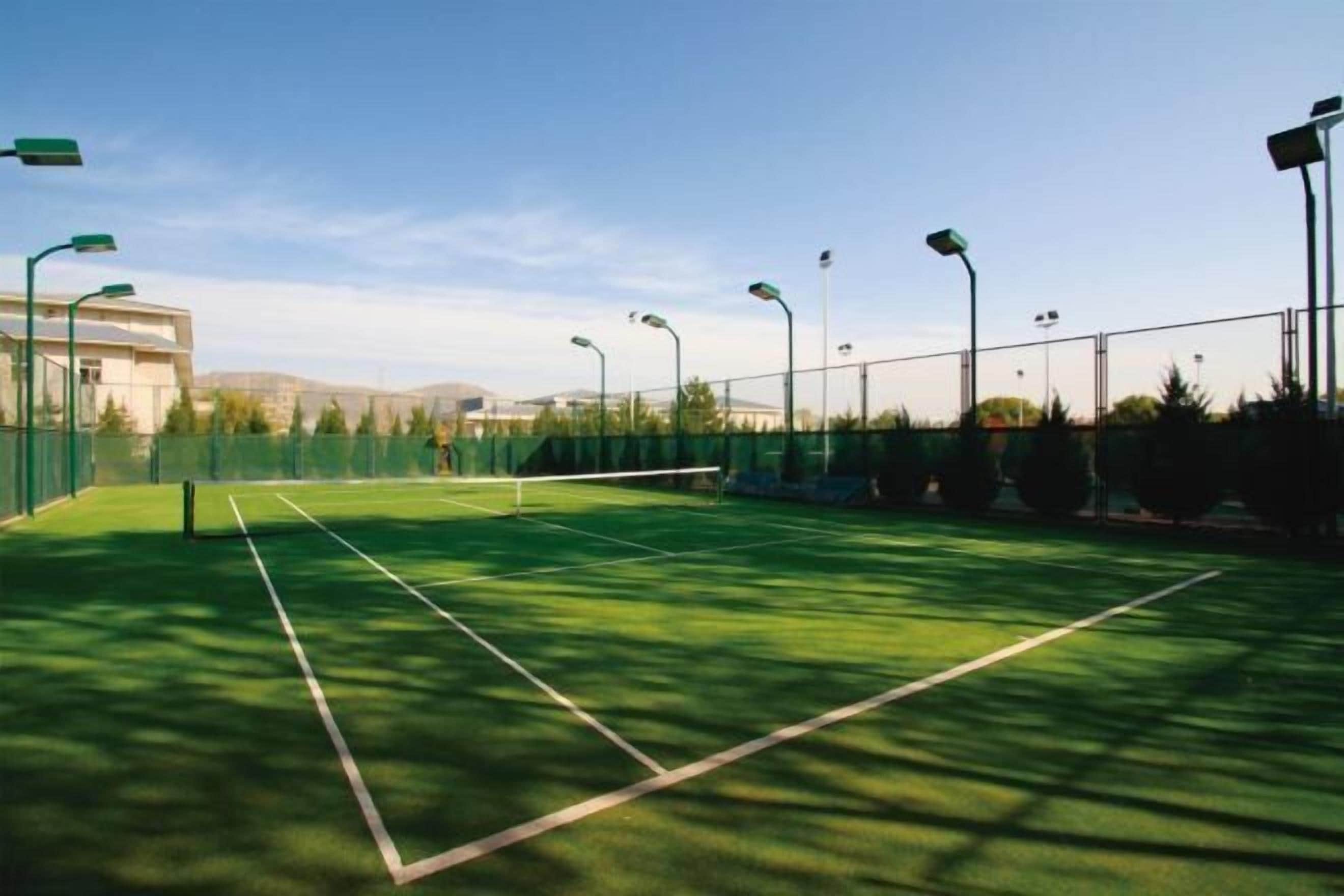 Ein Tennisplatz im Freien mit Netz, umgeben von einem Zaun und Straßenlaternen.