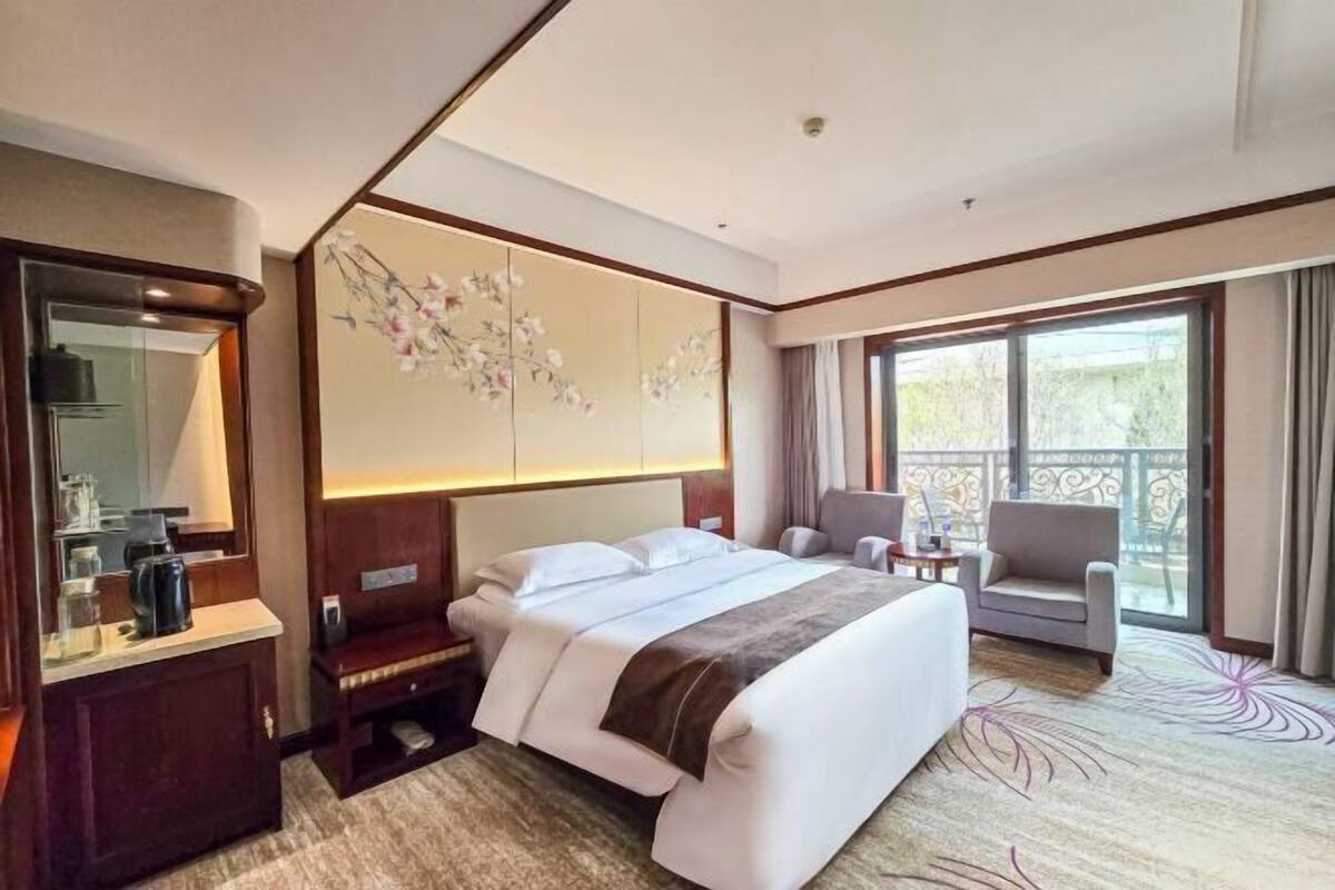Habitación de hotel con una cama grande, un escritorio con espejo, una silla y vistas al exterior a través de un ventanal amplio.