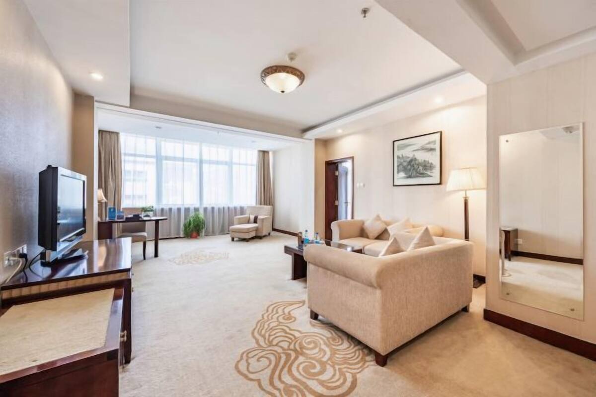Una habitación de hotel con sofá, televisor, escritorio, lámpara y un ventanal grande.