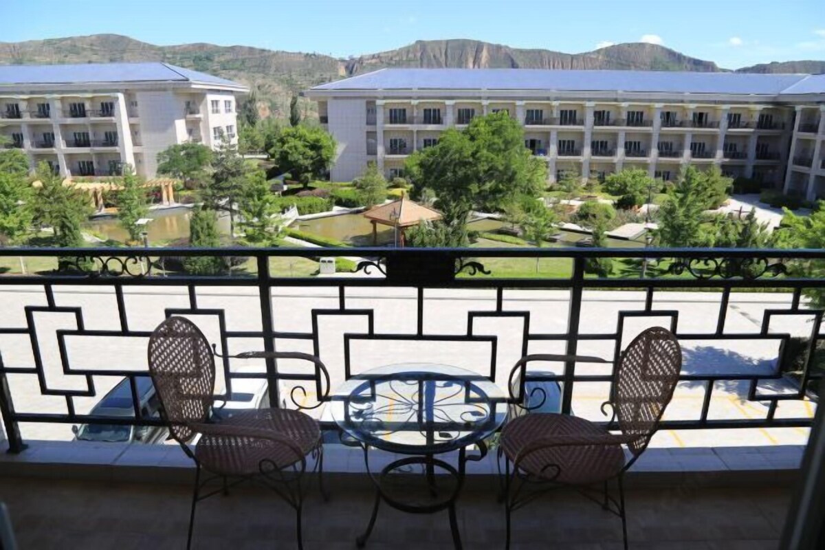 A balcony with two wicker chairs and a small table, overlooking a courtyard with a pool and buildings.