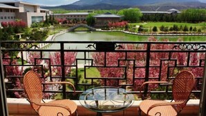 Un balcon agrémenté de fauteuils en osier et d’une table en verre, donnant sur un jardin aménagé avec un étang et, au loin, une montagne.