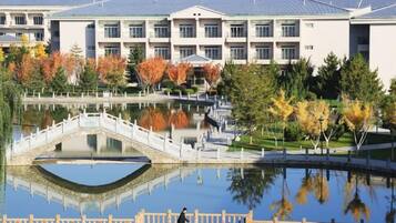 Un bâtiment avec une fontaine ou un bassin d’eau dont la surface réfléchit la lumière, un pont piétonnier et des arbres aux feuilles d’automne.
