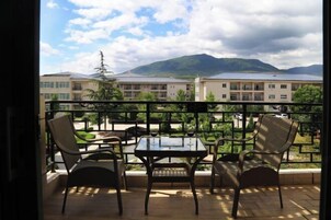 Un balcón con dos sillas de mimbre y una mesa de vidrio, con vistas a un edificio y montañas.