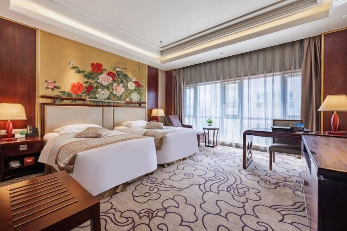 Habitación de hotel con dos camas, un escritorio, una silla, una mesita y un ventanal grande con cortinas.