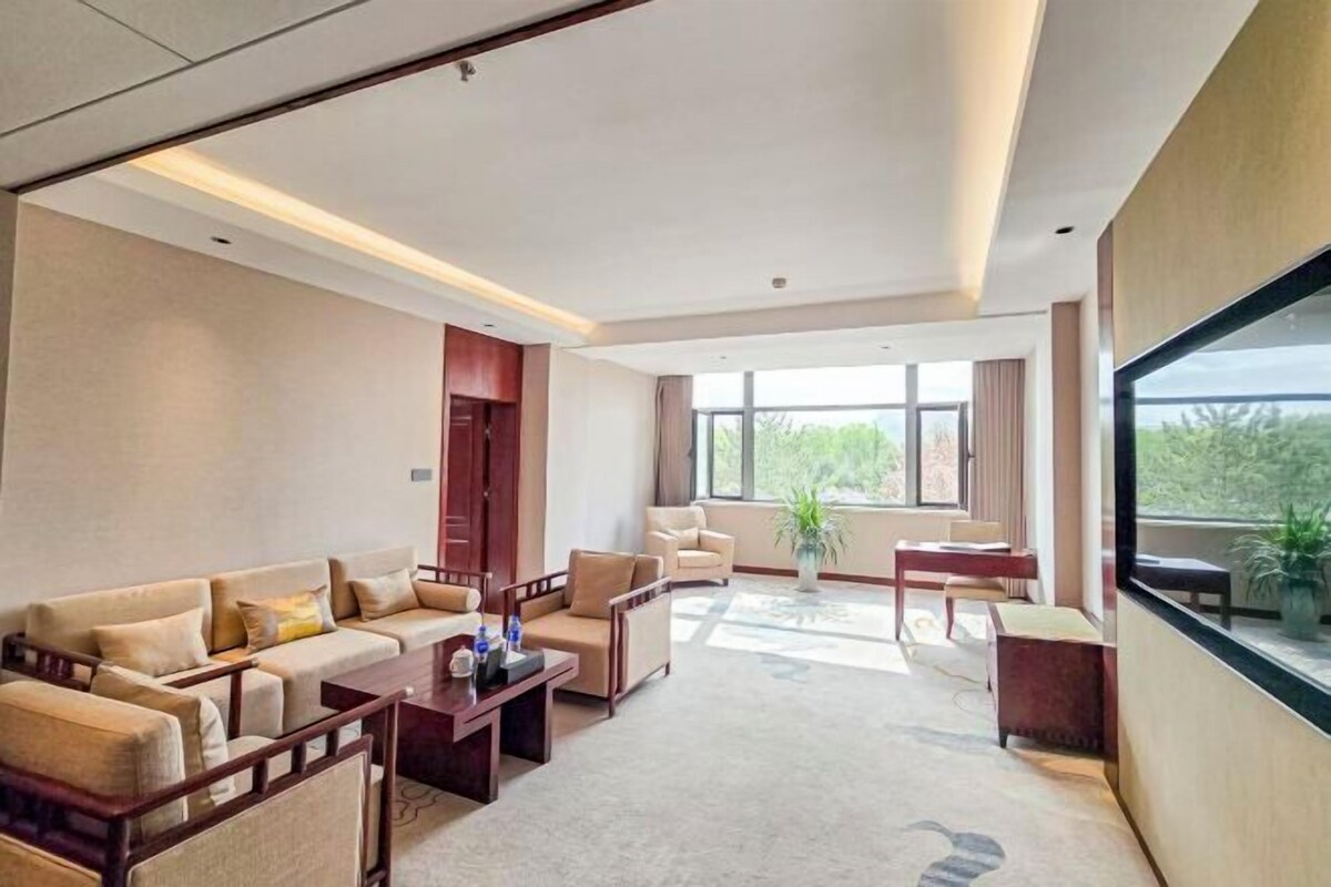 A spacious living area with a sofa set, a coffee table, and a dining table. There is a large window providing a view of greenery outside.