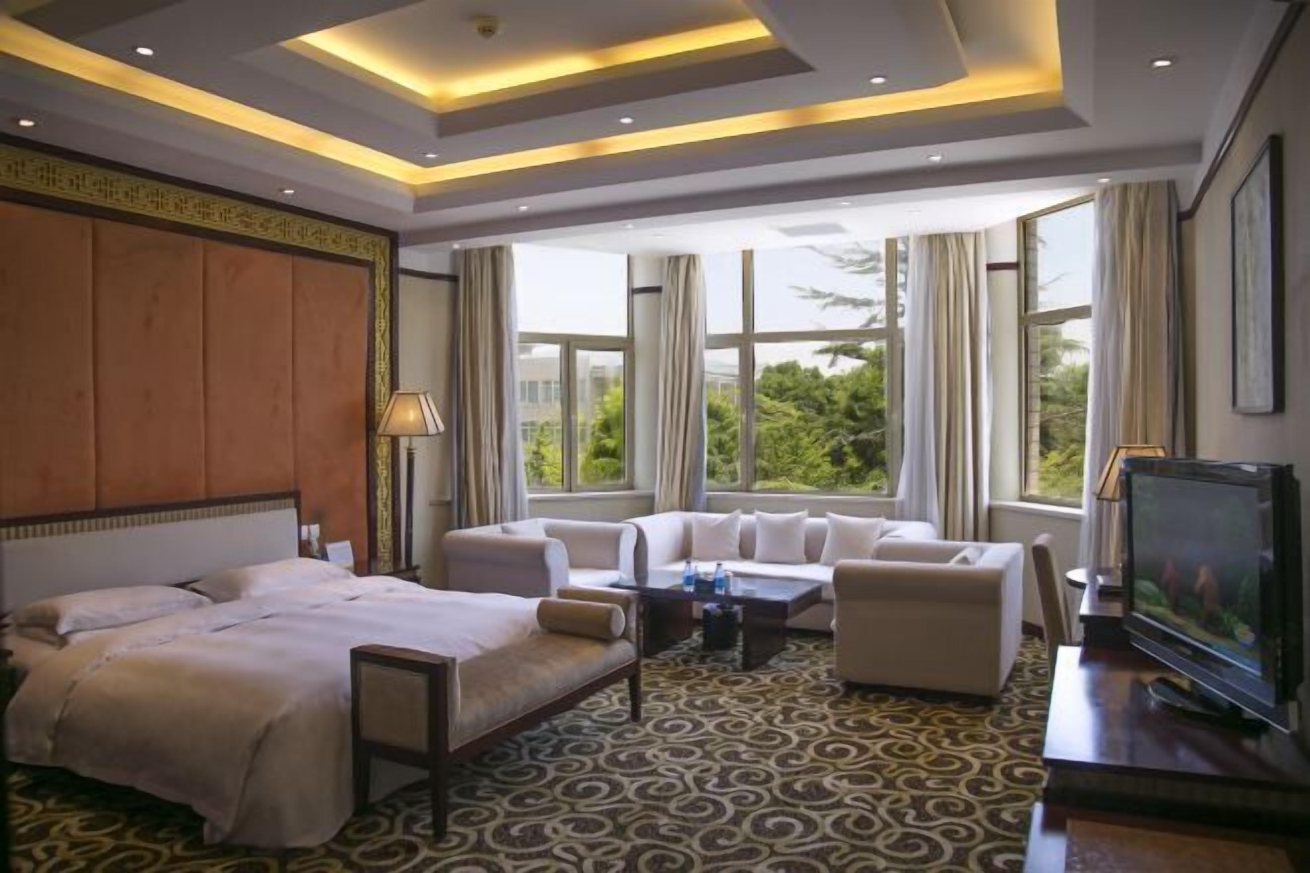 Una habitación de hotel con cama, sofá, televisor y ventanas amplias con cortinas.