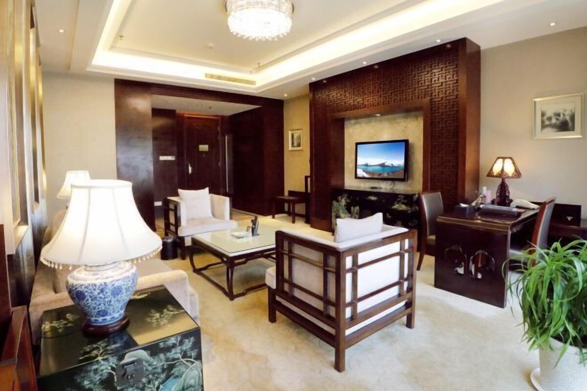 A modern hotel room with a large TV mounted on a brick wall, a sofa, a coffee table, and a desk with a lamp.