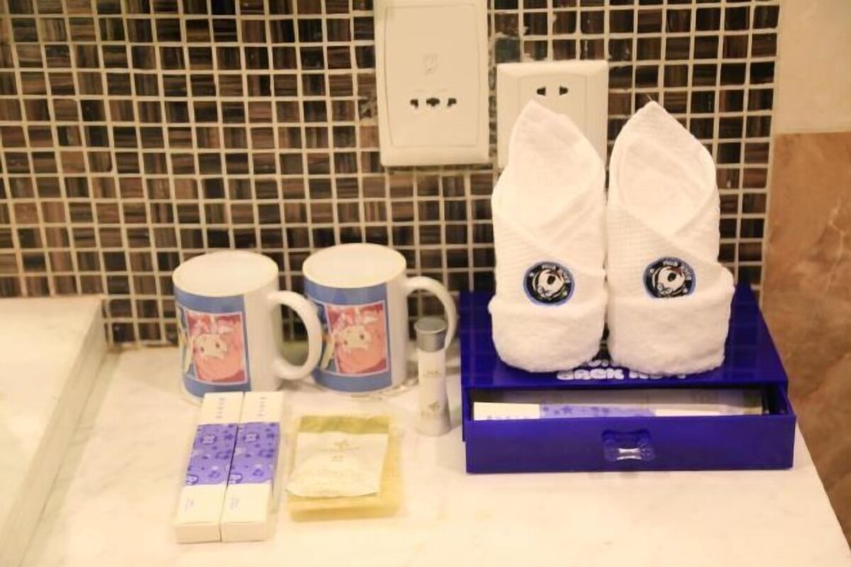 A bathroom with a tiled backsplash, a white electrical outlet, two white towels with cartoon eyes, two mugs with cartoon designs, and a blue box with a white item inside.