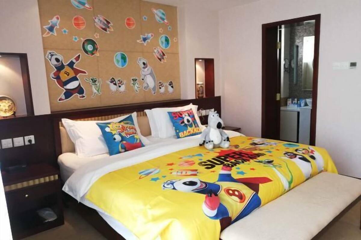 Una habitación de hotel con una cama y un edredón con motivos de superhéroes, una pared decorada con personajes de caricaturas y una mesita de noche con una lámpara.