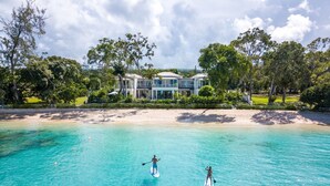 A beachfront property with a large house, clear turquoise water, and two people paddleboarding.