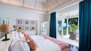 A bedroom with a large bed, a desk with a lamp, framed artwork on the wall, and a balcony with a view of the sea.