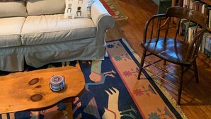 Ein gemütliches Wohnzimmer mit einer Couch, einem Holzstuhl, einem Couchtisch, einem Bücherregal und einem durch eine offene Tür einsehbaren Küchenbereich.