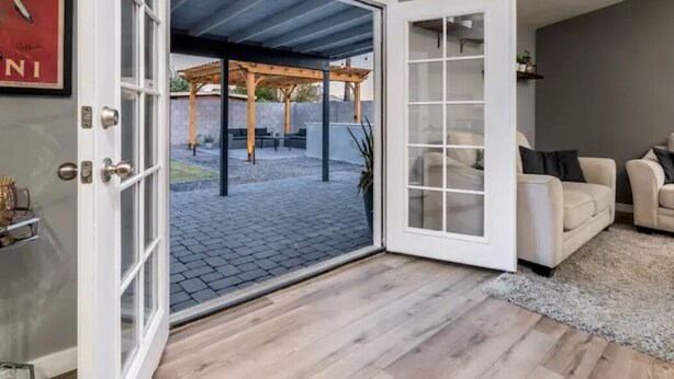 An open sliding glass door leading to an outdoor patio with a pergola, a beige sofa inside, and a framed picture on the wall.