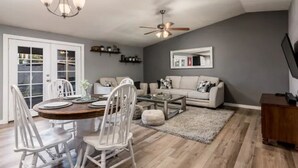 A spacious living room with a dining area, grey walls, wooden flooring, a ceiling fan, and a flat-screen TV mounted on the wall.