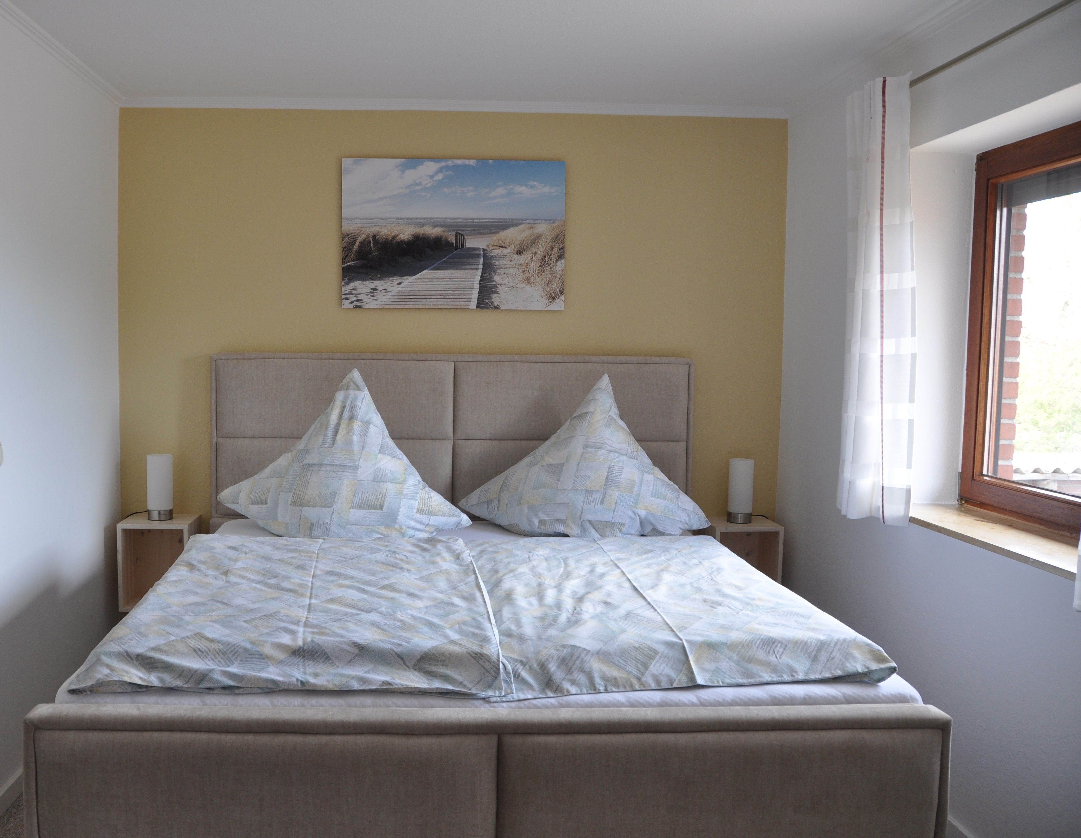 Ein Schlafzimmer mit einem Bett, zwei Nachttischen, einem Bild an der Wand und einem Fenster mit Vorhängen.