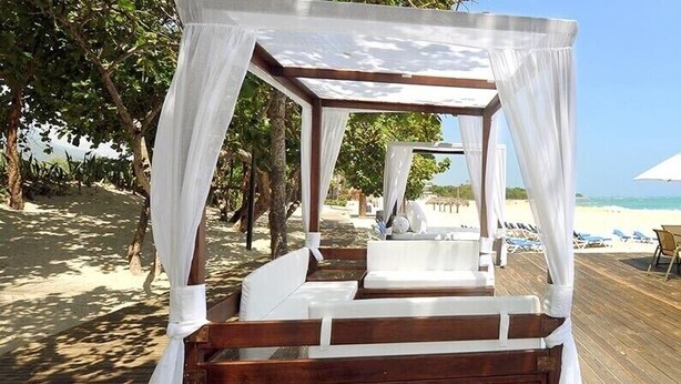 Eine Holzterrasse direkt am Strand mit einem Himmelbett, Strandstühlen und Sonnenschirmen.