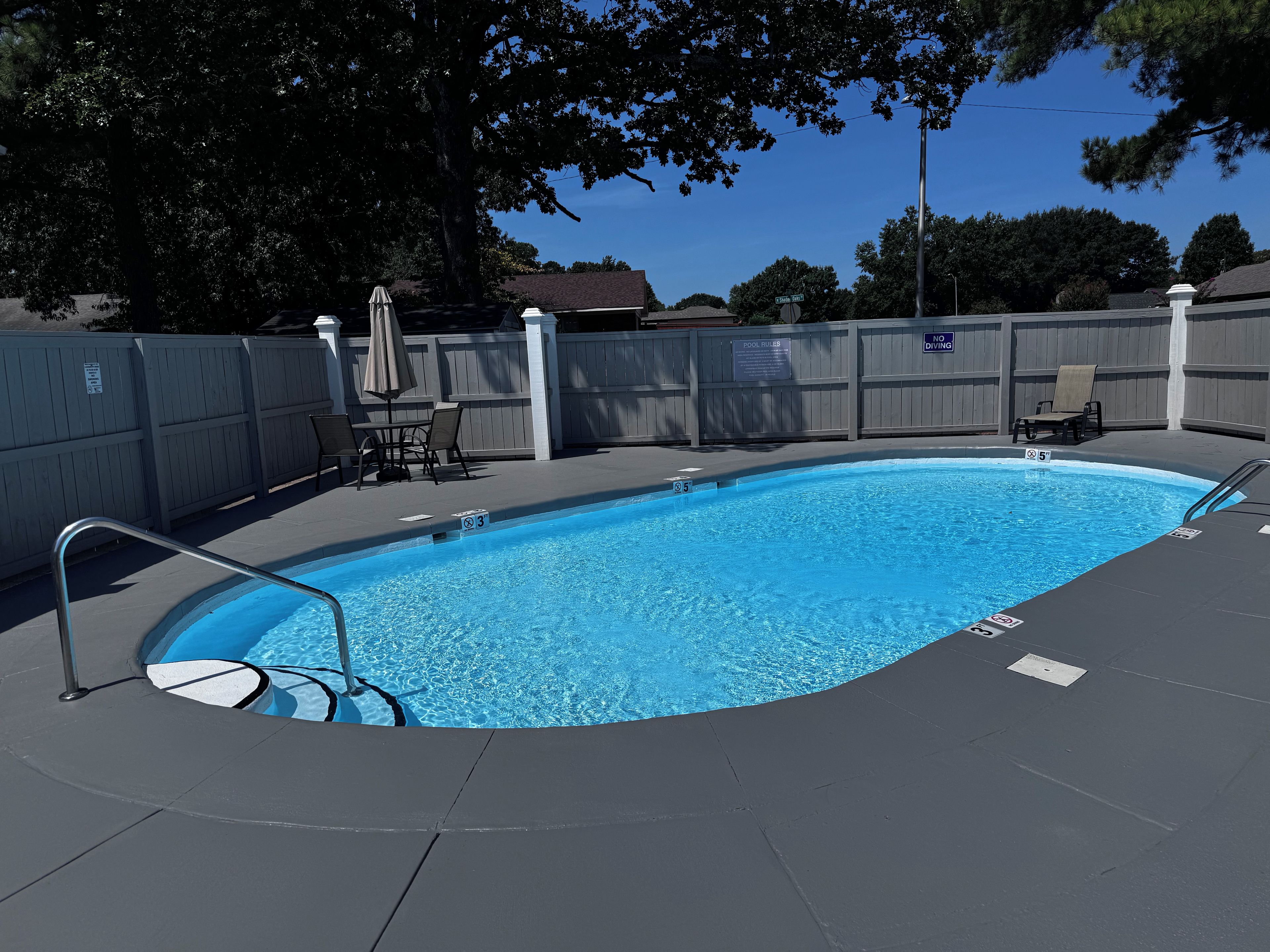 Una piscina rectangular con una escalera de metal, rodeada por una valla de madera y un área de descanso con sillas y una sombrilla.