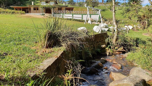 A rural setting with a small stream, grassy areas, and a building with a sloped roof in the background. - Sítio Pedacinho do Céu na Serra de SC (Aguas Mornas)