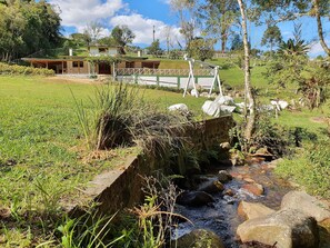 A rural setting with a small stream, grassy areas, and a building with a sloped roof in the background. - Sítio Pedacinho do Céu na Serra de SC (Aguas Mornas)