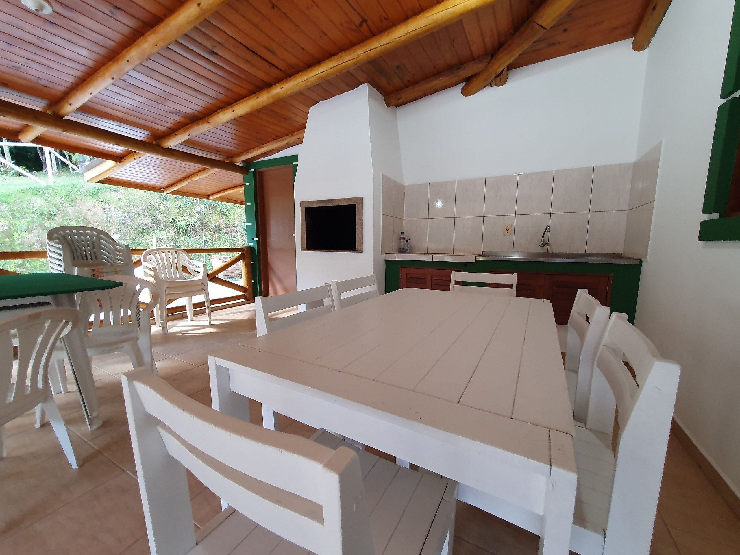 A covered outdoor dining area with a long table and chairs, a small kitchen with a sink and tiled backsplash, and a television mounted on the wall.