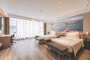 Habitación de hotel con dos camas, un escritorio, una silla, un banco, un ventanal grande con cortinas y un cuadro de una escena costera.