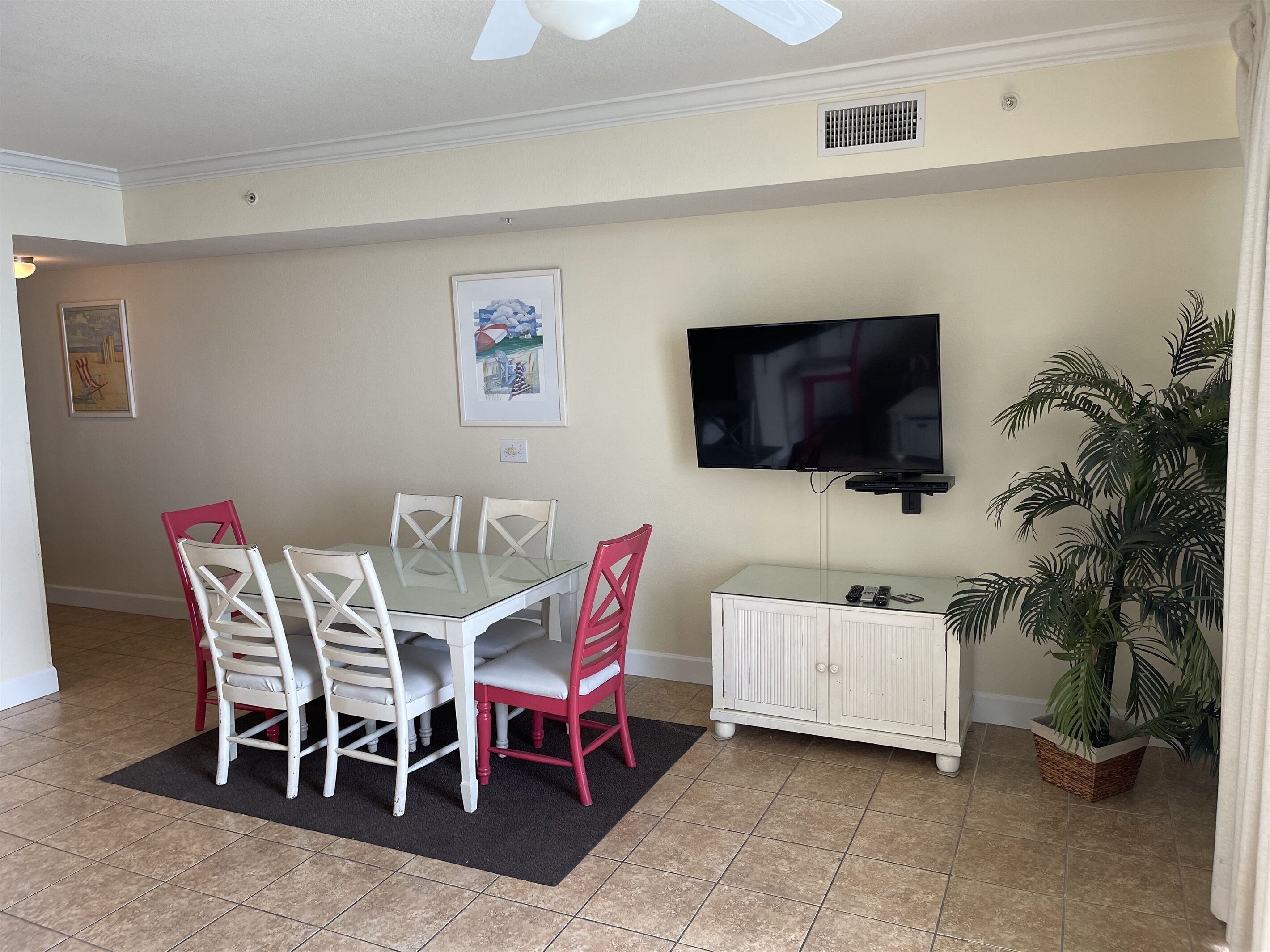A dining area with a glass table, white and red chairs, a TV mounted on the wall, a white cabinet with remote controls, and a potted plant.