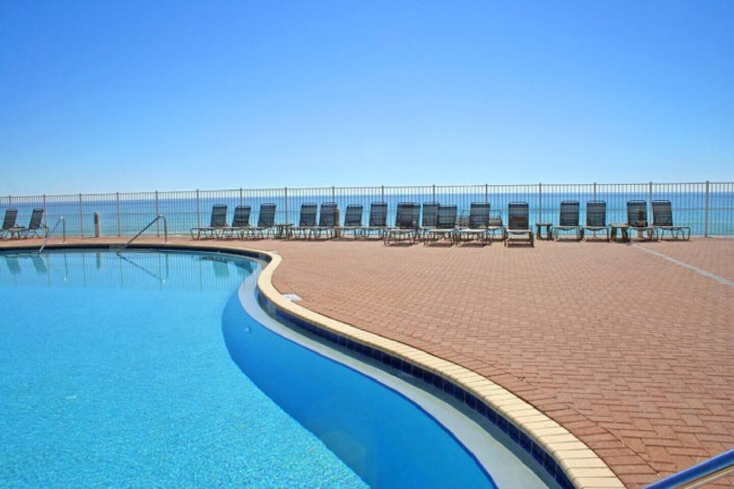 A swimming pool with a curved edge, surrounded by a brick patio and lounge chairs facing the ocean.
