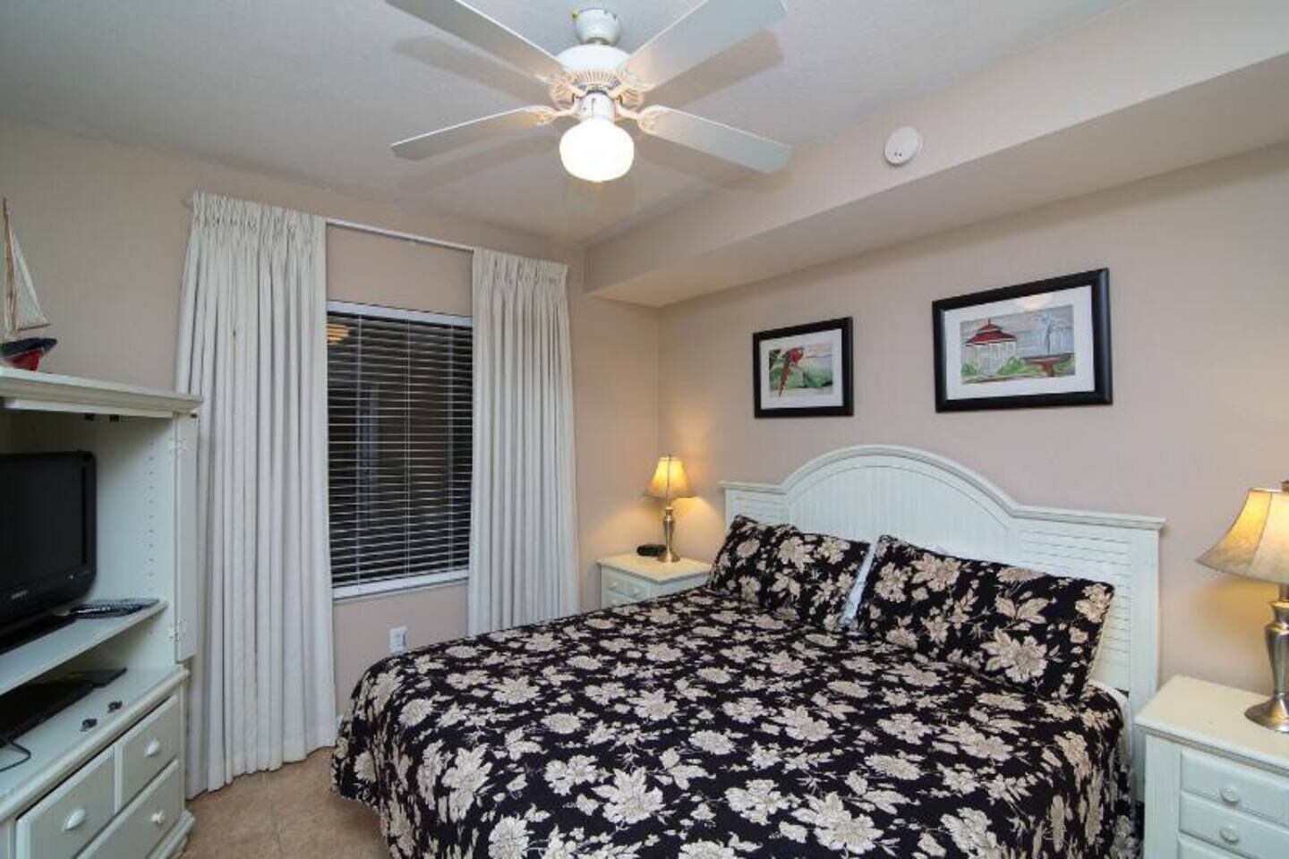 A bedroom with a bed, a ceiling fan, a TV, a lamp, and two framed pictures on the wall.