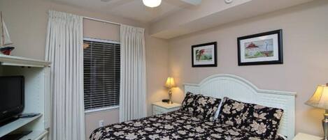 A bedroom with a bed, a ceiling fan, a TV, a lamp, and two framed pictures on the wall.