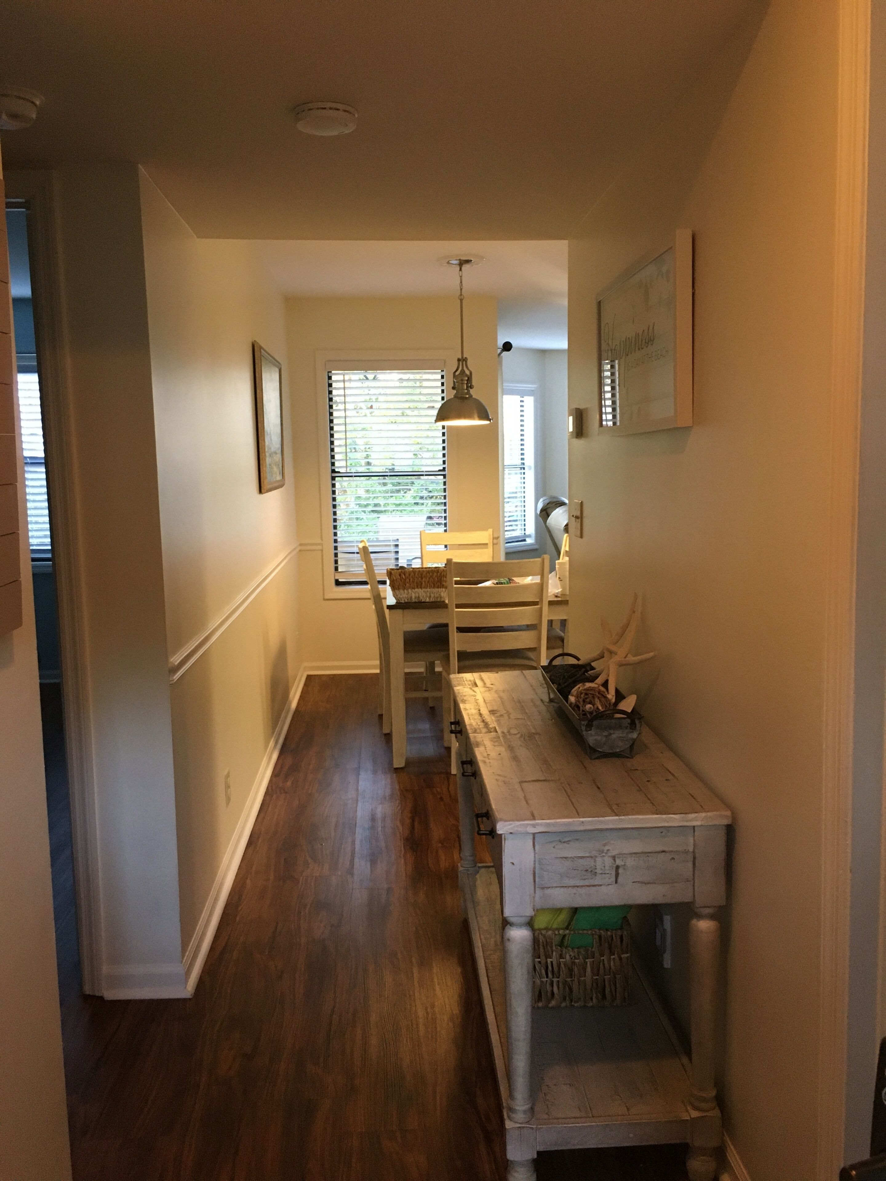 A narrow hallway with wooden flooring, a dining table with chairs, a hanging pendant light, and a small table with antlers and a basket.