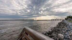 Uferlandschaft mit einer Holzpromenade, felsigem Ufer und einem entfernten Steg unter bewölktem Himmel.