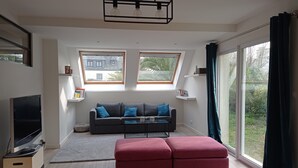 Una sala de estar moderna con un sofá gris, un reposapiés rojo y un televisor sobre un mueble bajo. Se ven dos ventanas con vistas a una casa y follaje.