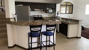 Une cuisine dotée de plans de travail en granit, de meubles de couleur sombre, d’un réfrigérateur en acier inoxydable, d’un four à micro-ondes et d’un évier. On y trouve également deux tabourets de bar avec des coussins bleus.