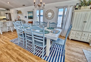 A dining area with a white table and blue chairs, a chandelier, a mirror, and a view of the ocean.