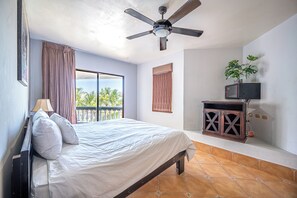 A bedroom with a bed, a ceiling fan, a window with curtains, a small TV on a cabinet, and a potted plant.