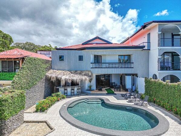A two-story house with a swimming pool, a thatched-roof structure, and a balcony.