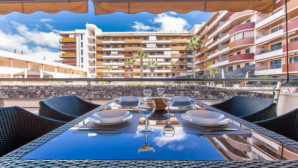 An outdoor dining area with a table set for two, overlooking a multi-story building with balconies and palm trees. - Balcon de Los Gigantes 2A016 poolview (Acantilados de Los Gigantes)