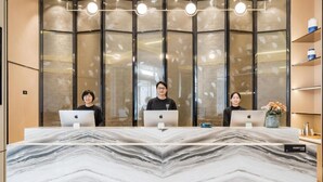 Un mostrador de recepción con tres personas sentadas detrás, cada una con una computadora portátil. El mostrador es de mármol con un diseño en forma de V. Encima del mostrador, hay varias lámparas colgantes. Al fondo, se observa una partición de vidrio con un motivo de hojas.