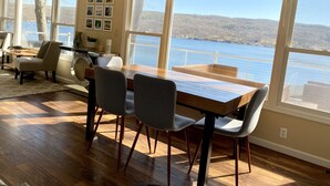 Un comedor con mesa y sillas de madera, ventanas amplias con vista a un lago, y techo con vigas a la vista.