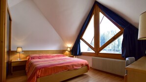 A bedroom with a bed, two bedside lamps, a window with curtains, a radiator, and a wooden desk.