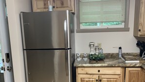 A kitchen with a stainless steel refrigerator, wooden cabinets, a window with blinds, and a countertop with various items. - Cottage in the Woods (Warfordsburg)