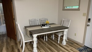 A dining area with a wooden table and four chairs, a small side table with a fruit bowl, and a window with blinds. - Cottage in the Woods (Warfordsburg)