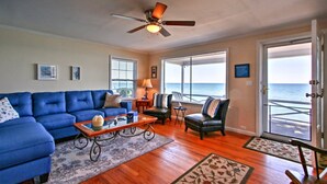 A living room with a blue sofa, a coffee table, and a ceiling fan. There are two windows with blinds, a door leading to a balcony, and a view of the ocean. - Fort Gratiot Lake House on Lake Huron w/ Patio & Deck! (Fort Gratiot Township)